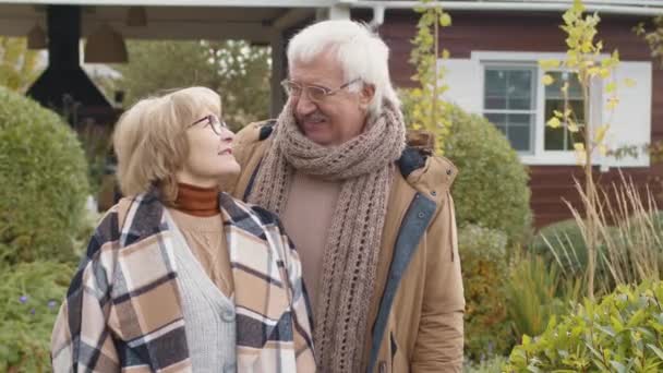Średnie ujęcie starszego mężczyzny rasy kaukaskiej i jego ukochanej pięknej żony w ciepłych ubraniach i okularach stojących w ogrodzie w pobliżu wiejskiego domu i patrzących przed kamerą - Materiał filmowy, wideo