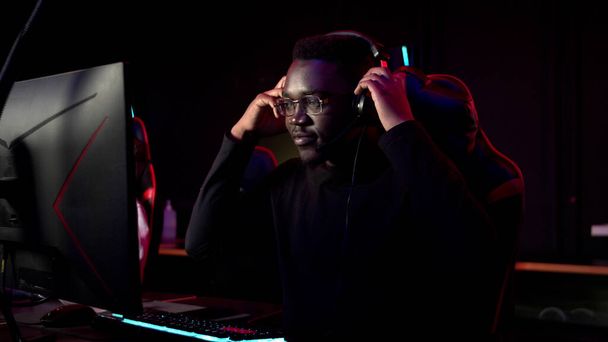 Un estudiante africano se sienta en una silla de juego en un club de computadoras, se pone auriculares, sonríe y se prepara para el juego - Foto, imagen