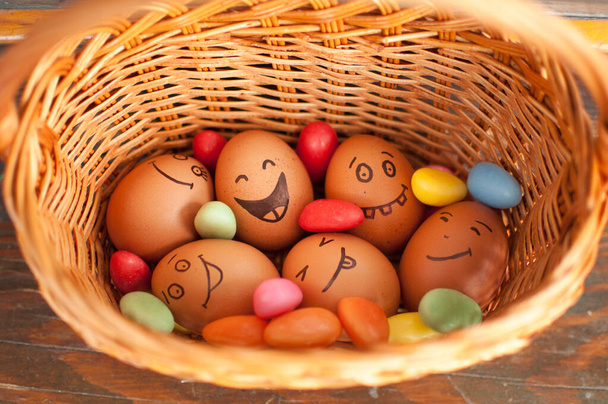 Στοίβα αυγών με ζωγραφισμένα στο χέρι πρόσωπα σε ψάθινο καλάθι με πολύχρωμα γλυκά, προετοιμασία του Πάσχα, ιδέες εορταστικής διάθεσης - Φωτογραφία, εικόνα