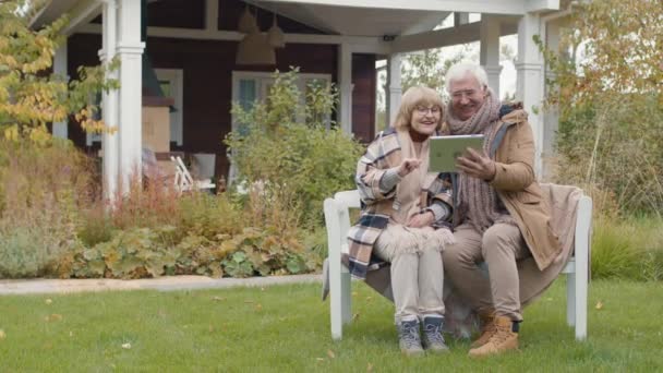 Široký záběr starších bělochů muž a žena v teplém oblečení sedí na lavičce na trávníku před jejich domem a mají video hovor pomocí digitálního tabletu - Záběry, video