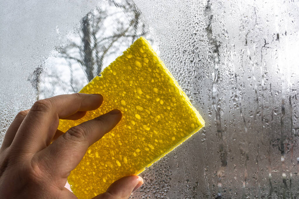 Un hombre limpia el vidrio empañado de una ventana de la humedad. En las manos de un paño amarillo. - Foto, Imagen