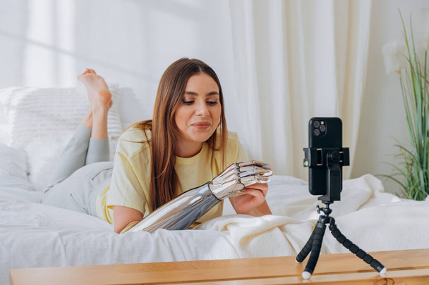 Женщина-блоггер с бионической протезной рукой ведет онлайн-трансляцию или записывает проблему или общается с друзьями по видеосвязи, пока лежит на кровати - Фото, изображение