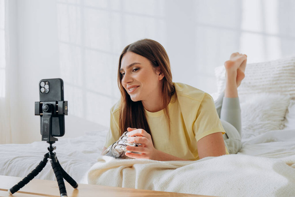 Bloggerin mit bionischer Armprothese führt eine Online-Sendung oder zeichnet ein Thema auf oder kommuniziert mit Freunden per Videoanruf, während sie auf einem Bett liegt - Foto, Bild