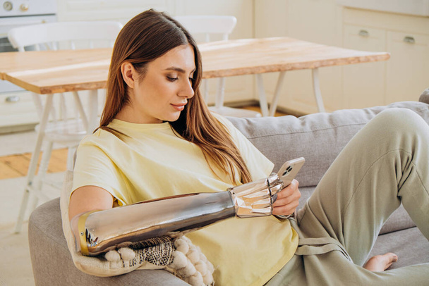 Una mujer feliz con un brazo protésico biónico se sienta en el sofá con un teléfono y se comunica en línea con amigos o navega por Internet o hace compras en línea - Foto, Imagen