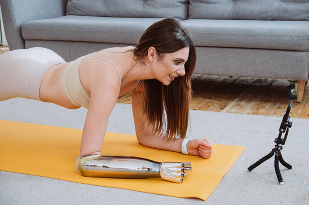 Uma jovem mulher com um braço protético biônico fica na prancha da casa em um tapete de ioga na frente de um telefone celular - Foto, Imagem