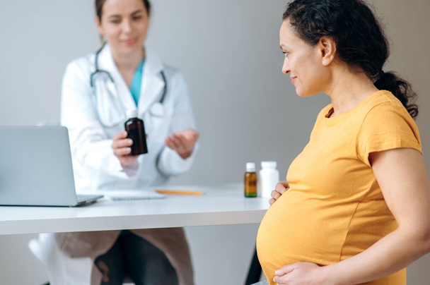 Doctor advises and prescribes medications for the pregnant woman. Mixed race, mature pregnant woman, sitting at the doctors appointment, undergoing medical prevention and treatment.Healthcare concept - Foto, afbeelding