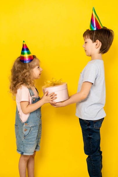 Little blonde girl accepts gift in pink box for her birthday from her brother, on a yellow background, children in caps, vertical photo. - Foto, Imagen