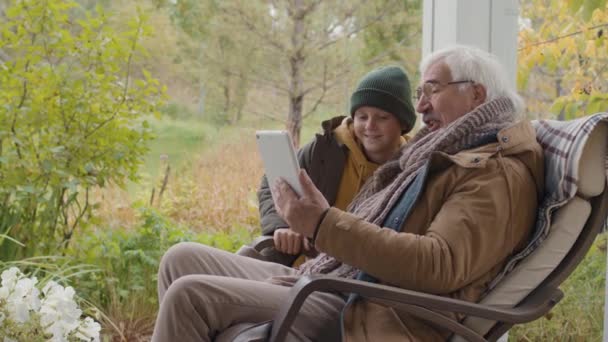 歳のロックダウンの白人男性に座っているロッキングチェア上のテラスと彼の十代の孫の両方を着て暖かい服を着てビデオ会話を使用してデジタルタブレット - 映像、動画