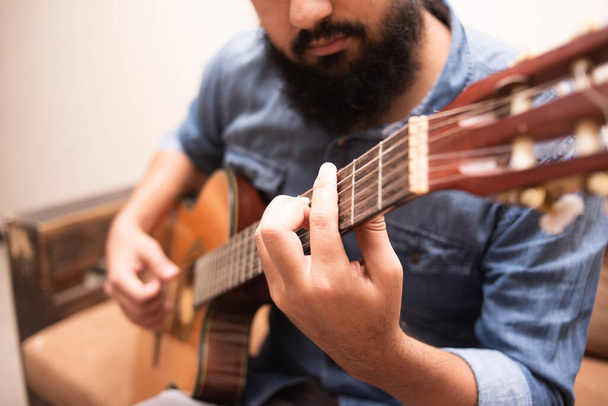 Homem vestido com roupas casuais tocando guitarra, sentado em uma poltrona em um quarto - Foto, Imagem