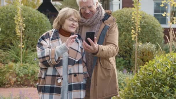 Μέτριο πλάνο του πρεσβύτερου καυκάσιου άνδρα και της συζύγου του που φορούν ζεστά καθημερινά ρούχα και γυαλιά που στέκονται στον κήπο κοντά στο σπίτι και έχουν βιντεοκλήση χρησιμοποιώντας τον άνθρωπο κινητό τηλέφωνο κρατώντας τα χέρια - Πλάνα, βίντεο