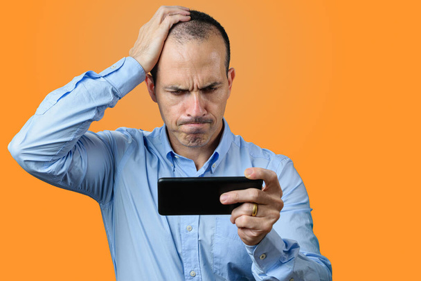 Uomo maturo in abito formale, guardando il suo smartphone, deluso e con la mano destra sulla testa. sfondo arancione. - Foto, immagini