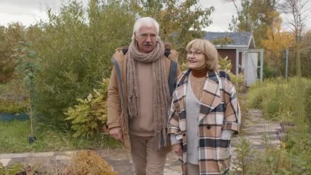 Foto mediana de viejo hombre caucásico con bigote y mujer, ambos con ropa casual cálida y anteojos caminando en un hermoso jardín y comunicándose - Metraje, vídeo