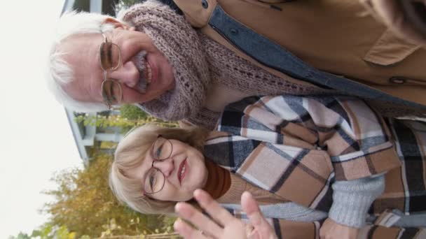 Verticale view shot van oude blanke vrouw en haar man dragen warme casual jassen en glazen buiten staan in de buurt van hun huis en het hebben van videogesprek met behulp van apparaat man in de hand - Video