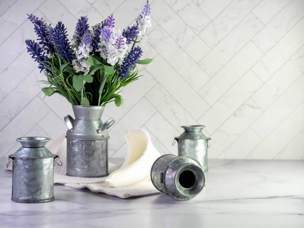 Rustikale Metallmilchdose gefüllt mit künstlichem violettem Lavendel und Flieder für ein einfaches Frühlingslandhausdekor mit Stillleben-Fotografie und einer Marmortheke mit Fischgrätfliesen-Hintergrund.   - Foto, Bild