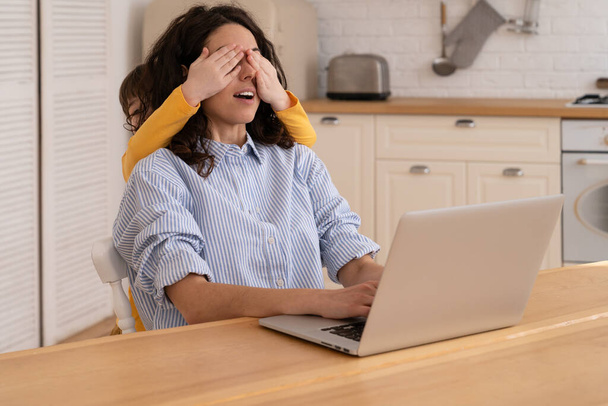 Мама працює на ноутбуці під час замикання, діти відволікаються від роботи, закриваючи очі матері.. - Фото, зображення