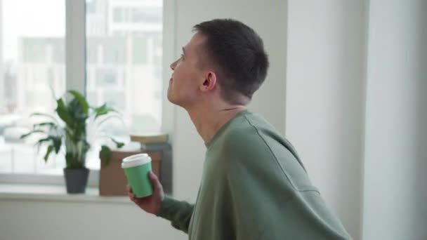Een man drinkt koffie uit een kartonnen beker bij het raam. - Video