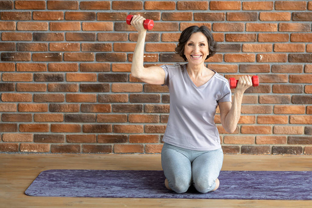 Happy smiling elderly woman fitness practitioner, sitting on fitness mat with dumbbells at home - Foto, Bild