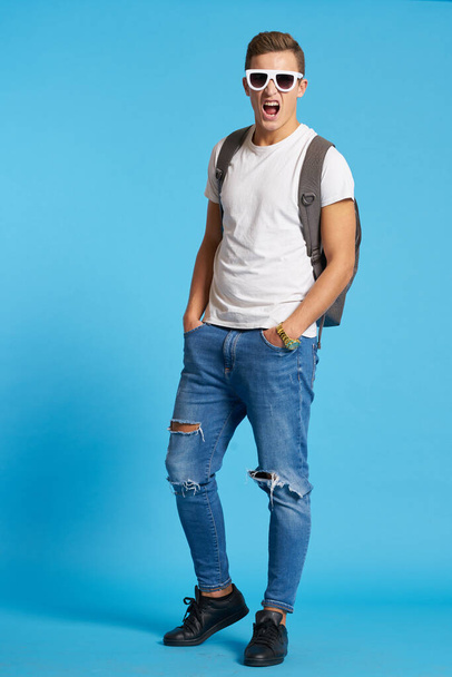 眼鏡とジーンズのスニーカーのバックパックを持つ男Tシャツ完全成長の青の背景 - 写真・画像