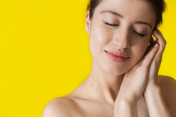 Caucasian freckled woman with bare shoulders is gesturing the sleep sign on a yellow studio wall advertising something - Photo, Image