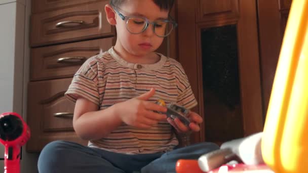 Caucásico niño pequeño con gafas sentado en el suelo y tratando de arreglar un juguete constructor - Imágenes, Vídeo