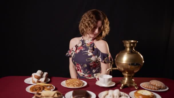 Chica con el pelo rizado y en el vestido de verano viene y se sienta a la mesa con deliciosos pasteles y samovar. Mujer marca en taza de agua para hacer té y bebe y disfruta sonriendo y cerrando los ojos - Imágenes, Vídeo