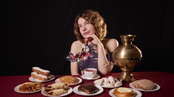 Una chica caucásica pensativa molesta se sienta en una mesa con un samovar y pasteles y piensa en algo y suspira y bebe té. La mujer está aburrida y sola, no puede encontrar un lugar - Metraje, vídeo