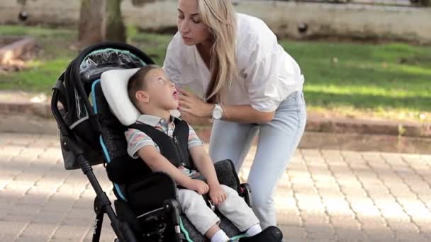 屋外で歩く幸せな愛の家族の母親と車椅子の瞬間に座っている障害のある息子の子供を見るのを助ける介護のビデオ。障害者介護の概念 - 映像、動画