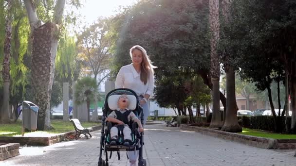 Βίντεο της οικογένειας μητέρα περπάτημα σε εξωτερικούς χώρους και φροντιστής βοήθεια ματιά σε ανάπηρο παιδί γιο κάθεται σε στιγμές αναπηρική καρέκλα. Έννοια φροντίδας αναπηρίας - Πλάνα, βίντεο