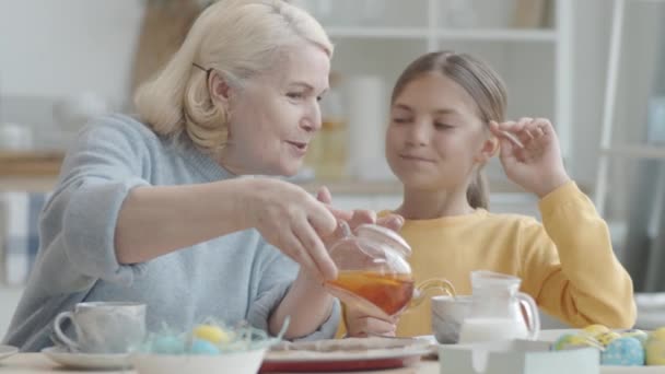 Mujer mayor sirviendo té en tazas, hablando con su nieta y escuchando galletas de conejo el día de Pascua - Imágenes, Vídeo