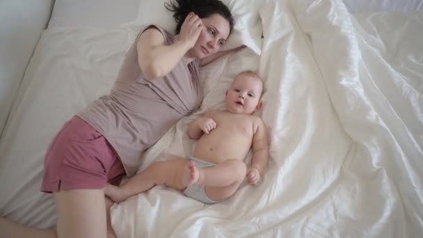ベッドの上にかわいい幼児の赤ちゃんの男の子と一緒に横たわっている幸せな若い母親は、腕の上に彼を保持し、午前中早く抱きかかえて遊んでいます。健康な子供,病院と幸せな母親の概念.保育園 - 映像、動画