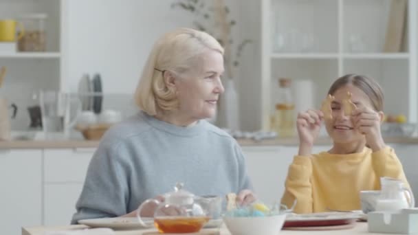 Zabawna dziewczynka trzyma dwa króliczki ciasteczka przed oczami, a następnie je jeść, siedząc razem z wesołą babcią przy stole w kuchni i jedząc kolację wielkanocną - Materiał filmowy, wideo