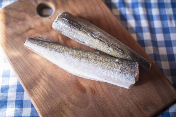 ωμό ψάρι σε ξύλινη σανίδα, υγιεινή χορτοφαγική τροφή, κατεψυγμένος μπακαλιάρος (Merluccius) σε πίνακα κουζίνας, μαγείρεμα ψαριών - Φωτογραφία, εικόνα