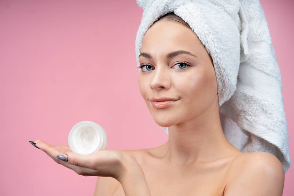 A young, beautiful, fresh beauty woman holds a jar of cream in her hands. female with towel on her head after spa, radiant face and skin care. On a solid background. - Fotoğraf, Görsel