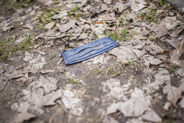 masques pour le visage Pollution, masque bleu sale recouvert de boue dans le parc sur le sol avec des feuilles sèches, masques jetables jetés par le coronavirus au printemps, anti-épidémiologique - Photo, image