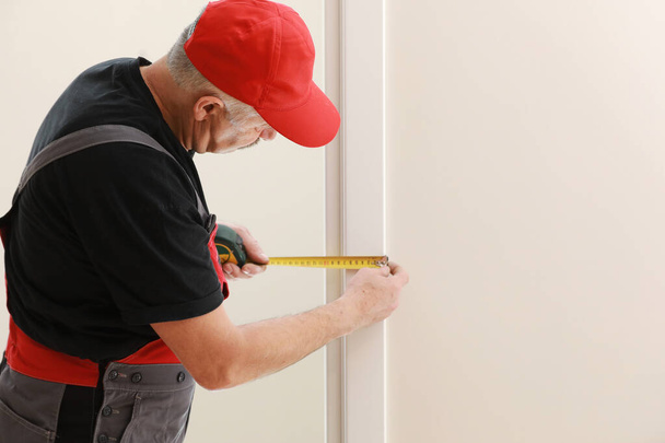 Pracovník v červené čepici a pracovním obleku s měřicí páskou. Muž instaluje dveře. Měřit pásku v rukou. Oprava funguje. Údržba v bytě - Fotografie, Obrázek
