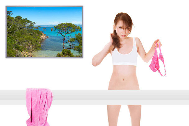 Jolie jeune femme portant un soutien-gorge et tenant une culotte rose à la main, elle se tient devant une photo d'une plage idyllique du sud, isolée sur fond de studio blanc - Photo, image