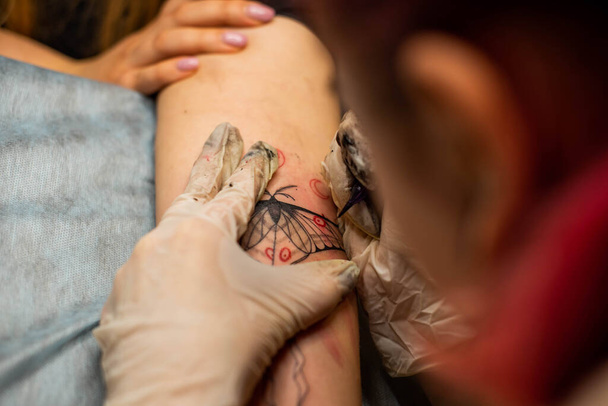 Il tatuaggio principale disegna la vernice rossa sul tatuaggio dei clienti. Tatuaggio artista in possesso di una macchina tatuaggio rosa in guanti sterili neri e lavorando sul tappeto blu professionale. - Foto, immagini
