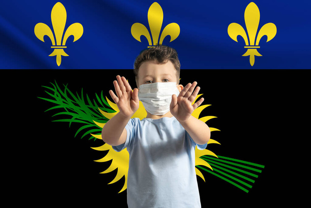 Μικρό λευκό αγόρι με προστατευτική μάσκα στο φόντο της σημαίας της Γουαδελούπης. Κάνει στοπ με τα χέρια του, μένει σπίτι Γουαδελούπη.. - Φωτογραφία, εικόνα