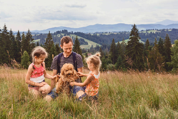 Ο μπαμπάς με δύο κόρες και ένα σκύλο σε ένα ξέφωτο σε ένα φόντο βουνών. Οικογένεια με ένα σκυλάκι σε πεζοπορία - Φωτογραφία, εικόνα