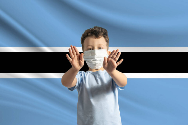 Μικρό λευκό αγόρι με προστατευτική μάσκα στο φόντο της σημαίας της Μποτσουάνα. Κάνει στοπ με τα χέρια του, μένει σπίτι Μποτσουάνα. - Φωτογραφία, εικόνα