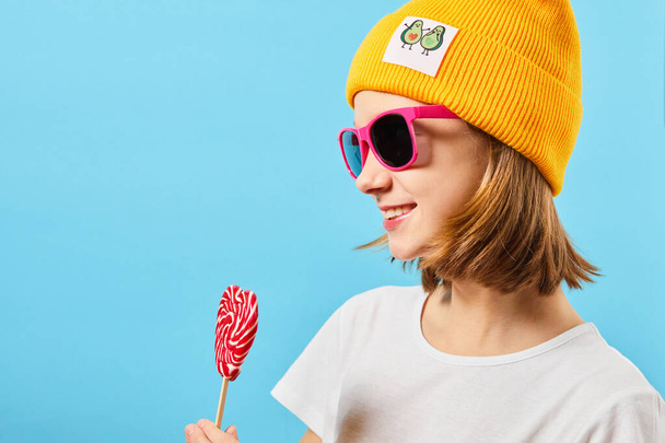 Hipster Teen girl in trendy glasses wearing knitted hat and holding lollipop. Fashionable woman with a candy near the face. - Zdjęcie, obraz