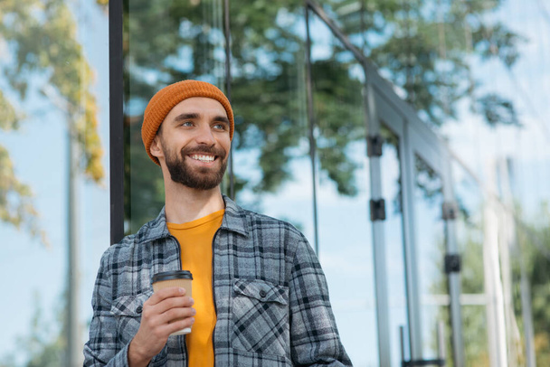 Knappe glimlachende man die koffiebeker vasthoudt terwijl hij buiten staat te kijken. Koffiepauze concept - Foto, afbeelding