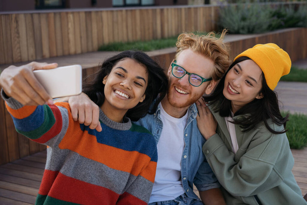 Group of emotional multiracial people using mobile phone taking selfies, smiling outdoors. African American woman influencer streaming video with happy friends. Positive lifestyle concept - Photo, Image