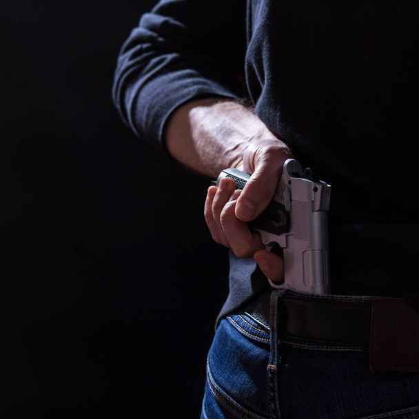 Mano masculina tomando un arma de su cinturón detrás de la espalda, sobre fondo negro. - Foto, imagen
