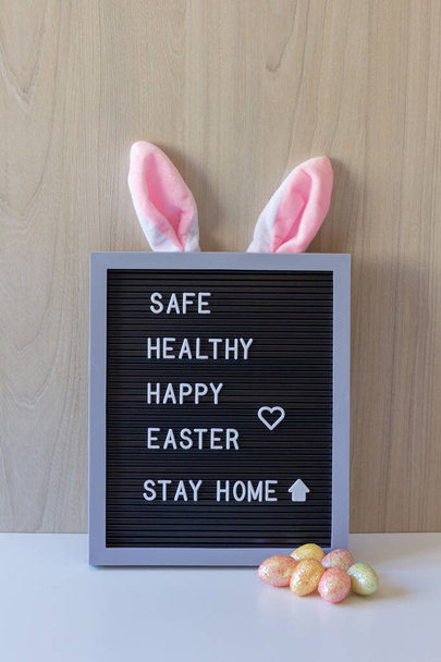 Koristeellinen merkki kanin korvilla ja värillisillä munilla pääsiäisviestin edessä: turvallinen, terve, onnellinen pääsiäinen, pysy kotona.. - Valokuva, kuva