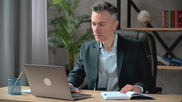 Успешный взрослый кавказский бизнесмен, адвокат или менеджер, в деловом костюме, работает на рабочем месте, пользуется ноутбуком, делает заметки, анализирует информацию, задумчиво смотрит в окно - Кадры, видео