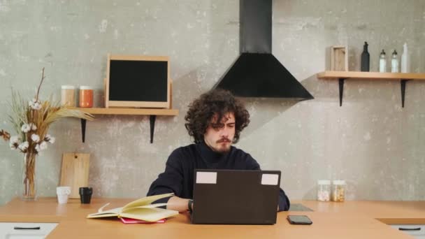 Vážné kudrnaté vlasy mladý muž pracující na počítači, zatímco sedí v kuchyni. - Záběry, video