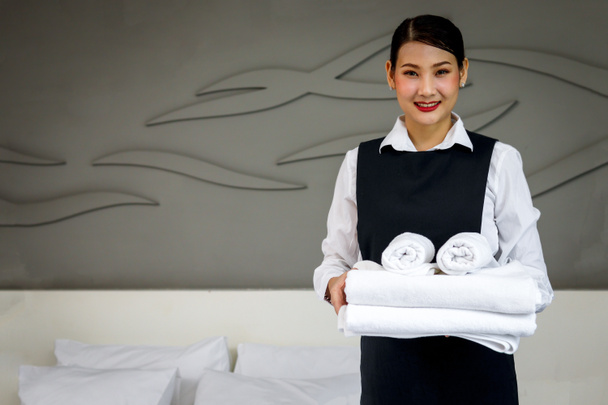 Room service maid cleaning and making bed hotel room concept, portrait of young beautiful Asian smiling female chambermaid holding stack of fresh white bath towels for making bed in hotel room. - Photo, Image