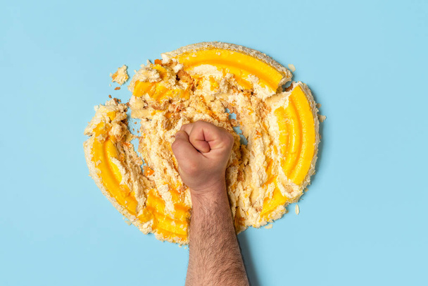 Above view with a man's fist smashing a cake. Concept for dieting with an orange whipped cream cake destroyed by a man, top view. - Photo, Image