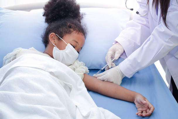 Врач вводит вакцину в руку афроамериканской девочки, лежащей на больничной койке. Чтобы укрепить иммунитет и защитить его от различных болезней. Концепция вакцины против COVID-19 - Фото, изображение